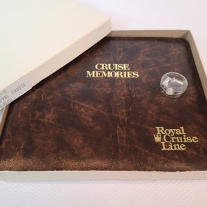 May include: A brown leather-bound photo album with the words "CRUISE MEMORIES" and "Royal Cruise Line" in gold lettering. The album is in a beige box with a clear plastic container of small objects. The album is open.