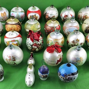 May include: A collection of Christmas ornaments with various designs, including snowmen, winter scenes, and festive patterns. The ornaments are primarily white and silver, with accents of red, blue, and gold. Some have bows and text.