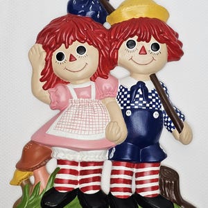 May include: A colorful ceramic wall hanging featuring two Raggedy Ann and Andy dolls. The dolls have red yarn hair, painted faces, and are dressed in pink, blue, and checkered outfits. They stand on green grass with a mushroom and a tree stump.
