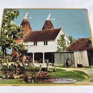 May include: A rectangular tin with a colorful illustration of a rural scene. The image features a white house with a brown roof, two tall, conical structures, and a pond. The tin has a gold-colored border.
