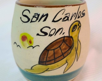 May include: Cream-colored ceramic tumbler with a hand-painted design. The tumbler features a yellow sea turtle, a yellow sun, and the text "San Carlos Son." in black. A blue stripe encircles the base.