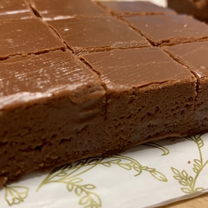 May include: A close-up of a chocolate fudge dessert cut into squares. The fudge is on a white paper with a gold leaf pattern.