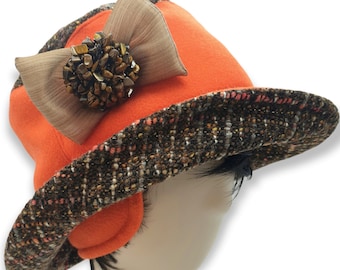May include: An orange cloche hat with a brown tweed brim and a decorative bow. The bow is tan and features a cluster of small, brown stones. The hat is a stylish accessory.