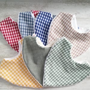 Peut inclure: Plusieurs bavoirs pour bébés de différentes couleurs, dont rouge, bleu, vert, jaune et beige. Chaque bavoir présente un motif vichy classique sur le devant et une doublure blanche et douce. Les bavoirs sont disposés en éventail.