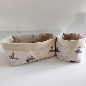 Peut inclure: Deux paniers de rangement en tissu beige. Les paniers ont un intérieur beige clair et un bord roulé. L'extérieur présente un motif répété d'une girafe de dessin animé dans un biplan. Les paniers sont de tailles différentes.