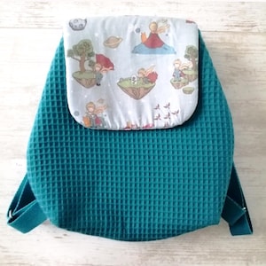 Peut inclure: Un sac à dos en tissu texturé gaufré bleu sarcelle avec un rabat gris clair orné d'un motif imprimé de petits personnages de dessin animé, d'îles flottantes, de planètes et d'oiseaux. Le sac possède des bretelles assorties bleu sarcelle.