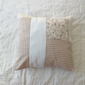 Peut inclure: Coussin carré décoratif avec un motif patchwork. Il présente des sections de vichy beige et blanc, de tissu blanc et un motif floral beige et marron. Le coussin est posé sur une surface blanche texturée.