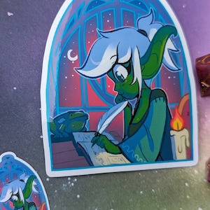 May include: A sticker featuring a green-skinned elf with white hair writing in a book with a quill. The elf is sitting in a window with a frog on the table next to them. The window is a stained glass design with a crescent moon in the upper left corner. The background is a gradient of pink, purple, and blue.