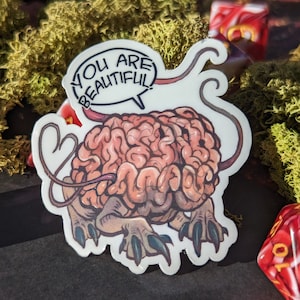May include: A sticker featuring a cartoon brain with a pink and gray color scheme. The brain has a tail and is holding a speech bubble that says "You are beautiful".