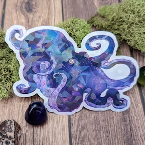 May include: A holographic sticker featuring a geometric octopus design in shades of purple, blue, and silver. The octopus has a starry sky pattern within its form. The sticker is on a wooden surface with moss and a dark purple stone.