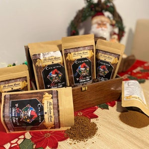 May include: Four brown paper bags with a Christmas theme. Each bag has a red and white label with the text "The Santa Barrel Coffee Company" and "Christmas Elf Pirate Brew Coffee". The bags are arranged in a wooden box with a red and white plaid fabric underneath. There is a pile of brown coffee grounds on the table.