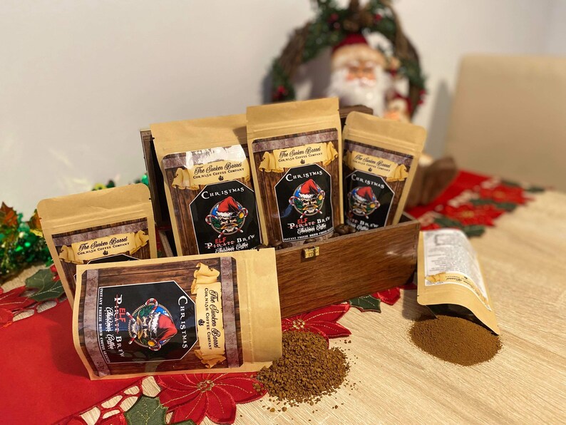 May include: Four brown paper bags with a Christmas theme. Each bag has a red and white label with the text "The Santa Barrel Coffee Company" and "Christmas Elf Pirate Brew Coffee". The bags are arranged in a wooden box with a red and white plaid fabric underneath. There is a pile of brown coffee grounds on the table.
