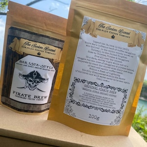 Może przedstawiać: Dwie złote torebki kawy z logo pirata i tekstem, który brzmi "The Sunken Barrel Cornish Coffee Company" i "Pirate Brew Legendary Celtic Coffee". Torebka po prawej stronie ma tekst, który brzmi "Original Pirate Brew Barista-Style Instant Coffee" i "100g".