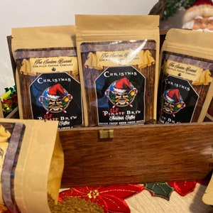 May include: A wooden box filled with five brown paper bags of coffee. Each bag has a festive Christmas design with a skull and crossbones and the words "Elf Pirate Brew Christmas Coffee".