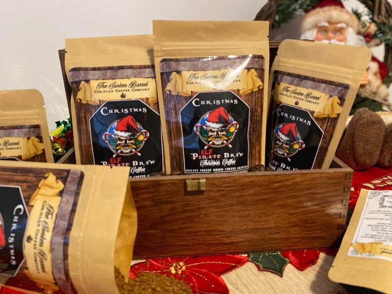May include: A wooden box filled with five brown paper bags of coffee. Each bag has a festive Christmas design with a skull and crossbones and the words "Elf Pirate Brew Christmas Coffee".