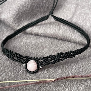 Może przedstawiać: Czarny naszyjnik typu choker wykonany techniką makramy z centralnie umieszczonym okrągłym, jasnoróżowym kamieniem. Opaska wykonana jest ze skomplikowanych węzłów sznurkowych i posiada regulowane zapięcie przesuwne do mocowania na szyi.