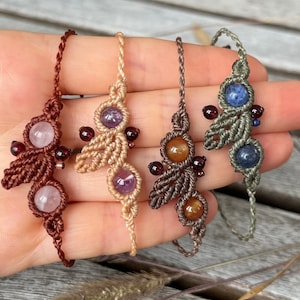 Könnte beinhalten: Vier Makramee-Armbänder mit Edelstein-Akzenten. Die Armbänder haben ein gewebtes Design in Braun-, Beige- und Grüntönen. Jedes Armband hat zwei größere Edelsteine und mehrere kleinere rote Edelsteine. Die Edelsteine sind rosa, lila und blau.