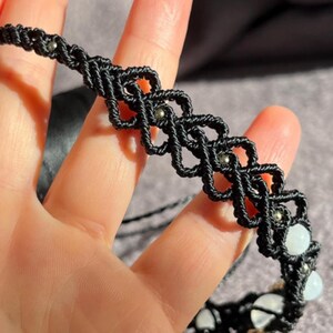 Puede incluir: Collar gargantilla de macram&eacute; negro con un dise&ntilde;o geom&eacute;trico, con peque&ntilde;as cuentas plateadas y detalles de piedras preciosas blancas. El collar hecho a mano se muestra sobre un fondo suave y neutro.