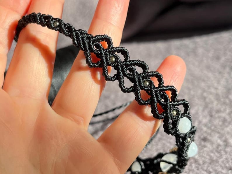 Puede incluir: Collar gargantilla de macram&eacute; negro con un dise&ntilde;o geom&eacute;trico, con peque&ntilde;as cuentas plateadas y detalles de piedras preciosas blancas. El collar hecho a mano se muestra sobre un fondo suave y neutro.