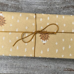 Puede incluir: Un paquete rectangular envuelto en papel marr&oacute;n con un patr&oacute;n repetido de erizos marrones y formas de &aacute;rboles blancos. El paquete est&aacute; atado con hilo dorado y un lazo.