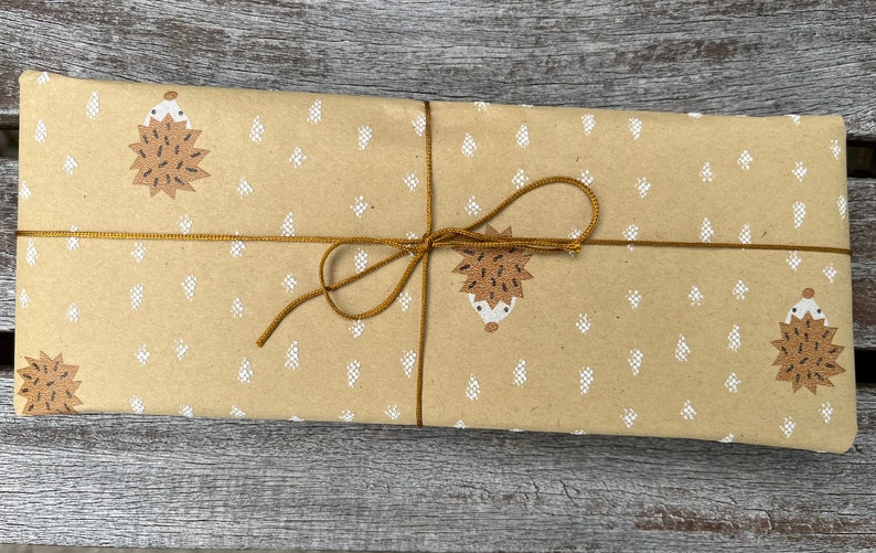 Puede incluir: Un paquete rectangular envuelto en papel marr&oacute;n con un patr&oacute;n repetido de erizos marrones y formas de &aacute;rboles blancos. El paquete est&aacute; atado con hilo dorado y un lazo.