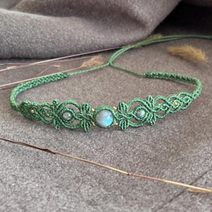 Puede incluir: Un collar gargantilla de macramé verde con una piedra preciosa central azul-gris y piedras preciosas más pequeñas a cada lado. El collar presenta intrincados nudos y pequeñas cuentas doradas. El collar se muestra sobre una superficie gris.