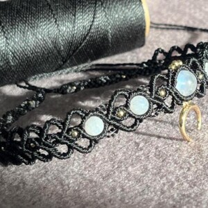 Puede incluir: Un collar choker de macram&eacute; negro con cuentas de piedra lunar y un dije de media luna dorada. El collar est&aacute; hecho con cord&oacute;n negro y presenta un patr&oacute;n decorativo. Una bobina de hilo negro est&aacute; en el fondo.