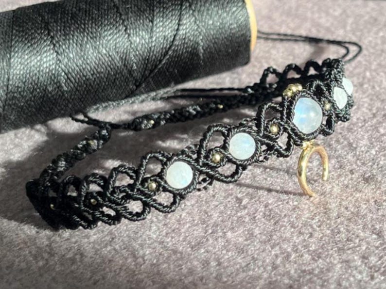 Puede incluir: Un collar choker de macram&eacute; negro con cuentas de piedra lunar y un dije de media luna dorada. El collar est&aacute; hecho con cord&oacute;n negro y presenta un patr&oacute;n decorativo. Una bobina de hilo negro est&aacute; en el fondo.