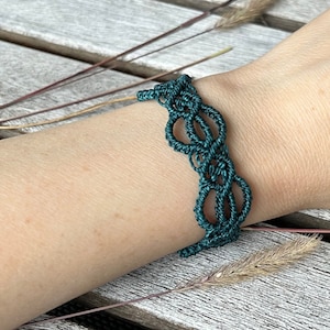May include: A dark green macrame bracelet with a woven pattern. The bracelet is on a person's wrist.