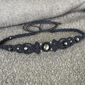 Può includere: Una collana girocollo in macramè blu scuro con perline grigio scuro e accenti floreali. La collana presenta un motivo intrecciato con piccole perline bianche e una chiusura con lacci. Il girocollo è esposto su una superficie grigia strutturata.