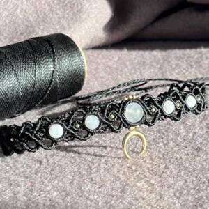 Puede incluir: Collar gargantilla de macram&eacute; negro con piedras redondas de color claro y un dije de media luna dorada. El collar se muestra junto a una bobina de hilo negro sobre una superficie p&uacute;rpura. Joya hecha a mano.