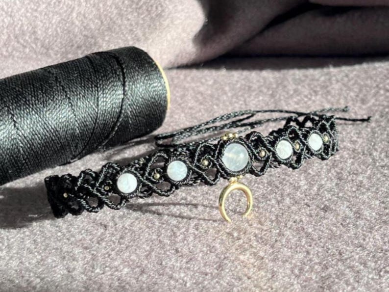 Puede incluir: Collar gargantilla de macram&eacute; negro con piedras redondas de color claro y un dije de media luna dorada. El collar se muestra junto a una bobina de hilo negro sobre una superficie p&uacute;rpura. Joya hecha a mano.