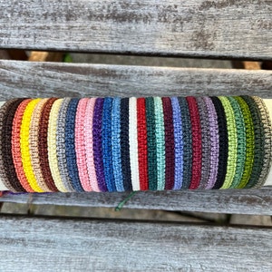 Könnte beinhalten: Eine Sammlung farbenfroher Makramee-Armbänder in verschiedenen Farbtönen, darunter Braun, Gelb, Rosa, Blau und Grün. Die Armbänder werden auf einer neutralen zylindrischen Oberfläche präsentiert, die ihre gewebte Textur und lebendigen Farben hervorhebt. Die Armbänder sind etwa 1,3 cm breit.