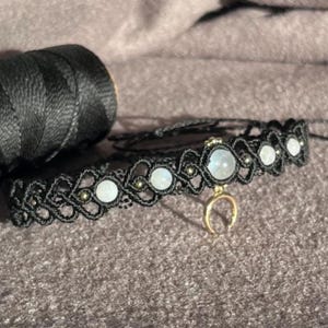 Puede incluir: Un collar choker de macram&eacute; negro con cuentas de piedra lunar y un dije de media luna dorada. Una bobina de hilo negro est&aacute; en el fondo. El collar est&aacute; hecho a mano y presenta un dise&ntilde;o tejido.