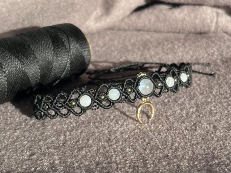Puede incluir: Un collar choker de macram&eacute; negro con cuentas de piedra lunar y un dije de media luna dorada. Una bobina de hilo negro est&aacute; en el fondo. El collar est&aacute; hecho a mano y presenta un dise&ntilde;o tejido.