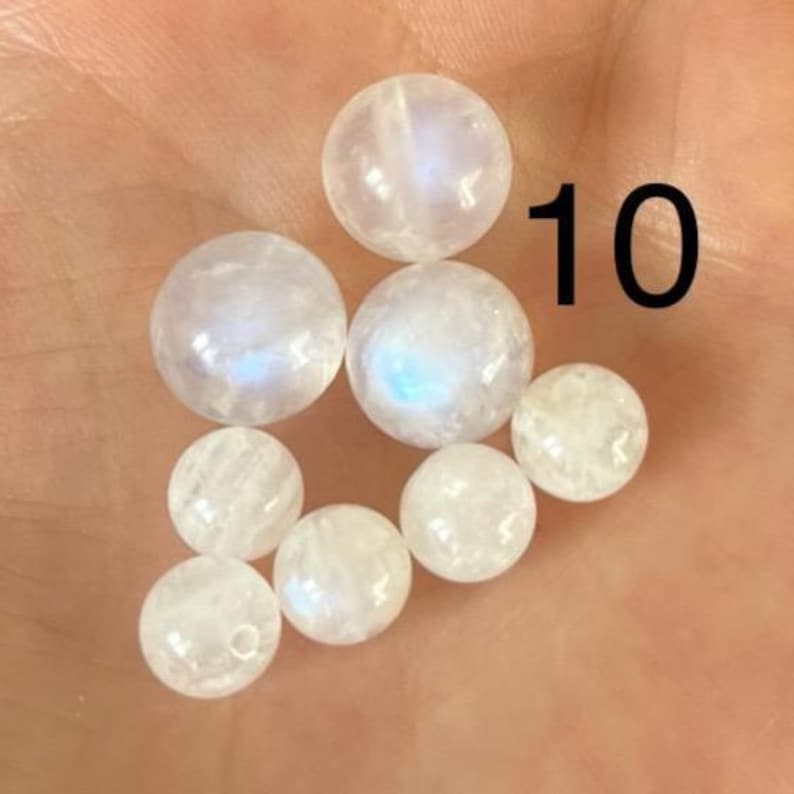 Puede incluir: Una colecci&oacute;n de nueve cuentas redondas de piedra lunar transl&uacute;cida. Las cuentas var&iacute;an en tama&ntilde;o y muestran una sutil iridiscencia, con toques de azul y blanco. El n&uacute;mero "10" est&aacute; impreso en negro en el fondo.