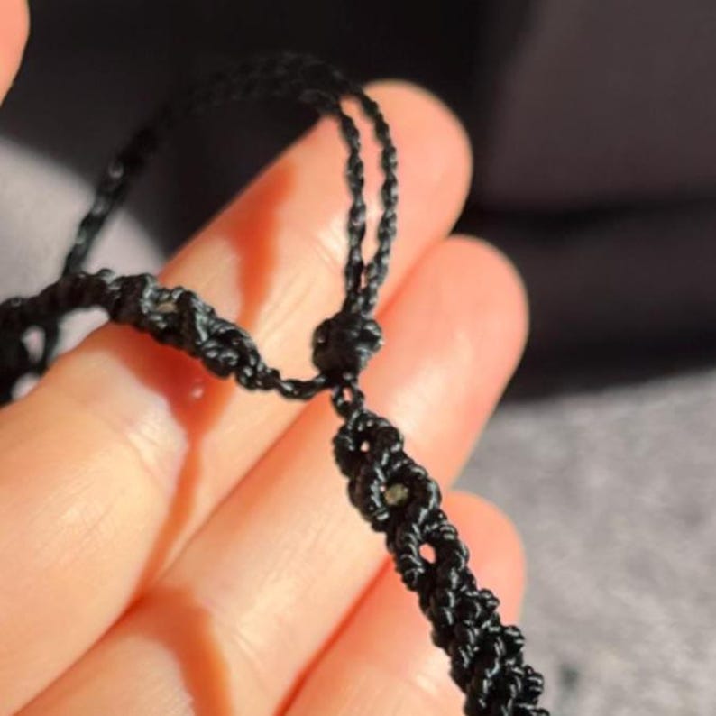 Puede incluir: Primer plano de un collar de macram&eacute; negro. El collar presenta un dise&ntilde;o tejido con un cierre de nudo corredizo. El cord&oacute;n de macram&eacute; est&aacute; anudado para crear un patr&oacute;n texturizado. El fondo est&aacute; borroso.