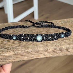 May include: A black macrame choker necklace featuring a central round iridescent gemstone flanked by two smaller round iridescent gemstones, accented with small silver-toned beads woven into the intricate black cord pattern with adjustable ties.