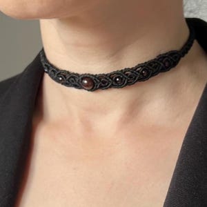 Può includere: Una collana girocollo in macramè nero con una perla centrale marrone scuro e perline più piccole. Il design intrecciato è visibile, creando un aspetto strutturato. La collana è indossata attorno al collo, mostrando la maestria artigianale e lo stile.