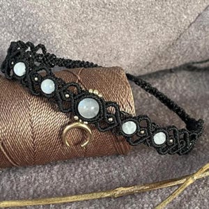 May include: Black macramé choker necklace with moonstone beads and a gold-coloured crescent moon charm. The necklace has small gold-coloured beads and is displayed on a spool of brown thread.