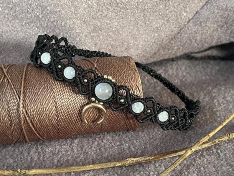 Puede incluir: Collar gargantilla de macram&eacute; negro con cuentas de piedra lunar y un dije de media luna dorada. El collar est&aacute; adornado con peque&ntilde;as cuentas doradas y se exhibe sobre una bobina de hilo marr&oacute;n.