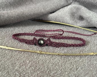 Puede incluir: Una gargantilla de macramé de color morado oscuro con una piedra preciosa redonda de color gris oscuro en el centro con un brillo sutil. El cordón está anudado en un patrón decorativo, con dos cuentas negras más pequeñas y lazos ajustables.