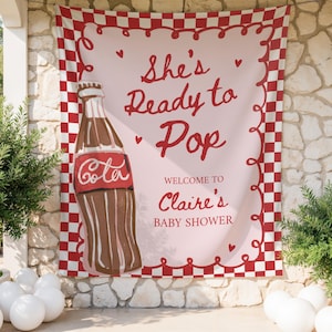 May include: A baby shower banner featuring a vintage soda bottle illustration. The banner reads "She's Ready to Pop" and "Welcome to Claire's Baby Shower" in red script on a pink background, framed by a red and white checkered border.