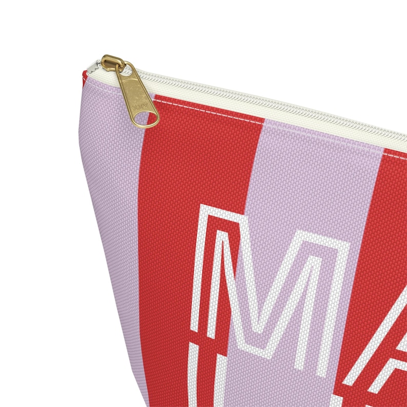 May include: A red and lavender striped cosmetic bag with a gold zipper. The bag features the word "MA" in white block letters. The bag is made of a durable fabric and is designed to hold makeup and other beauty products.