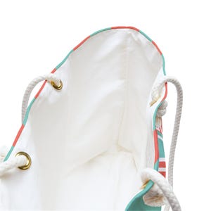 May include: White tote bag with a colorful design. The bag features a white interior, rope handles, and gold-colored grommets. The exterior has a pattern of red, white, and teal stripes.