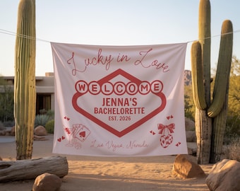 Pode incluir: Uma faixa branca com texto e gráficos vermelhos, lendo "Lucky in Love WELCOME JENNA'S BACHELORETTE EST. 2026 Las Vegas, Nevada". A faixa está pendurada ao ar livre com cactos ao fundo.
