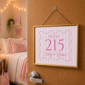 May include: A rectangular sign with a gold frame hanging by a rope on a wooden door. The sign features a light pink and white vertical striped background with a pink scalloped border, the text ROOM 215, and the names Ruby & Grace in pink script.