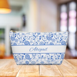 May include: A white cosmetic bag featuring a blue paisley pattern and the name "Abigail" in script. The bag has a gold zip and a white band across the centre. The paisley design includes floral and teardrop shapes.