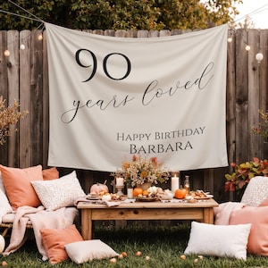 May include: A festive outdoor birthday scene with a banner that reads "90 years loved, Happy Birthday Barbara." The setting includes a wooden table with pumpkins, candles, and food, surrounded by pillows and a wooden fence.