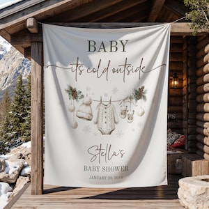 May include: A white banner with the text "BABY its cold outside" in brown script. The banner features illustrations of baby clothes and snowflakes. The banner also includes the text "Stella's BABY SHOWER JANUARY 20, 2023".